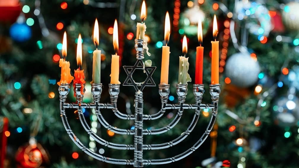 A lit menorah on the eighth night of Hanukkah with a Christmas tree in the background.